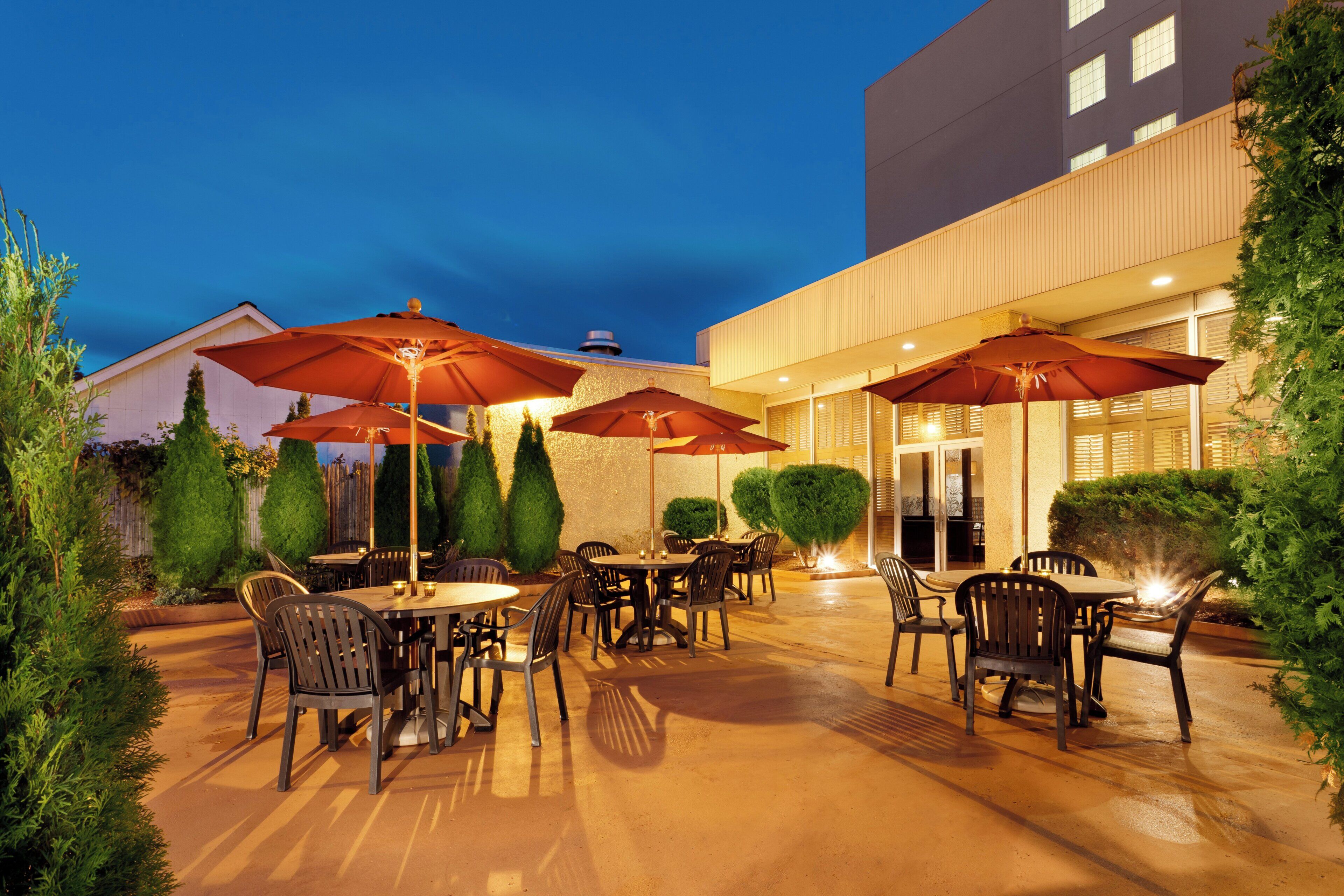 An outdoor patio area with tables and chairs, surrounded by greenery and umbrellas.
