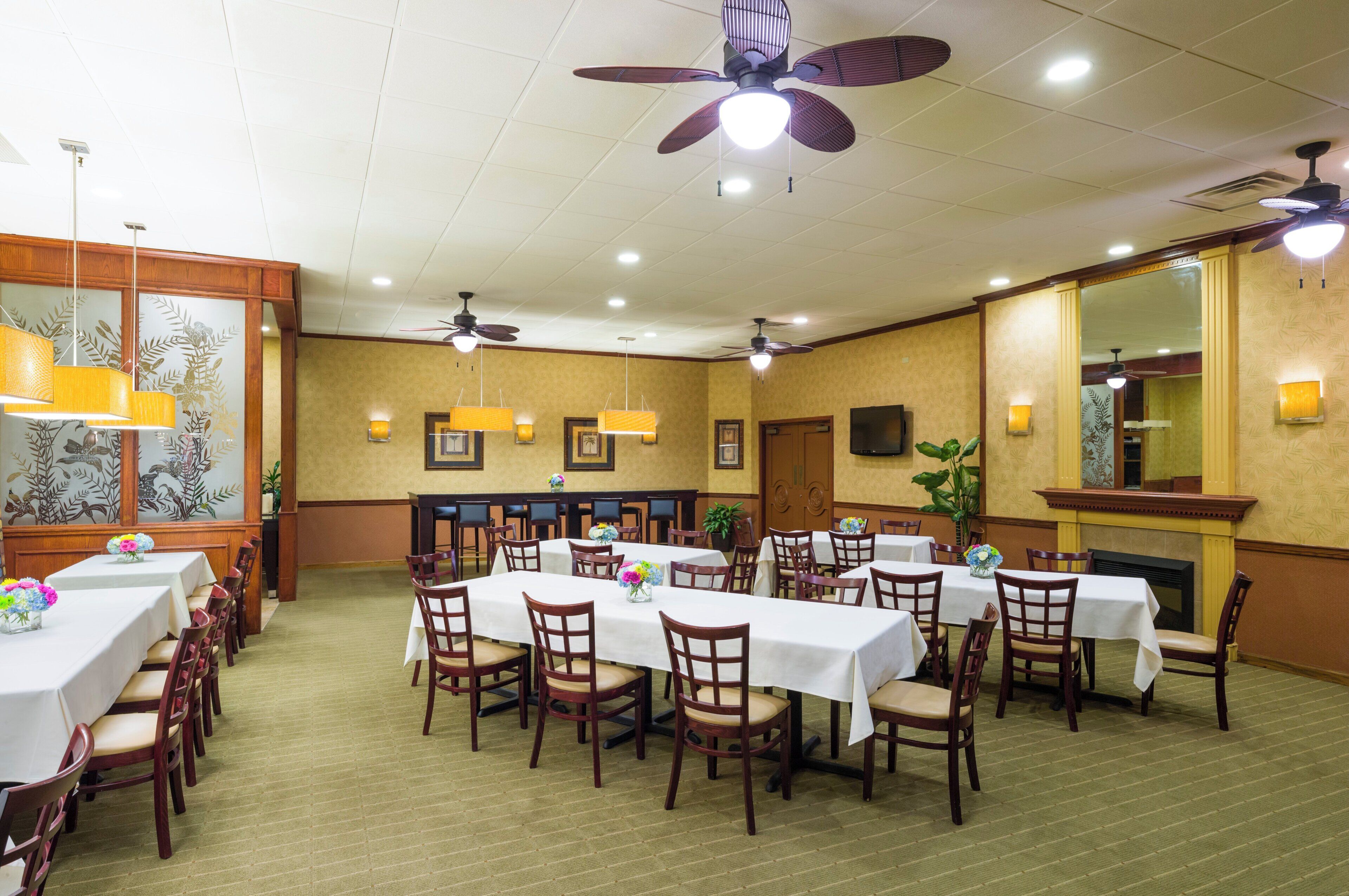 A hotel dining area with multiple tables and chairs, ceiling fans, and wall-mounted lighting.