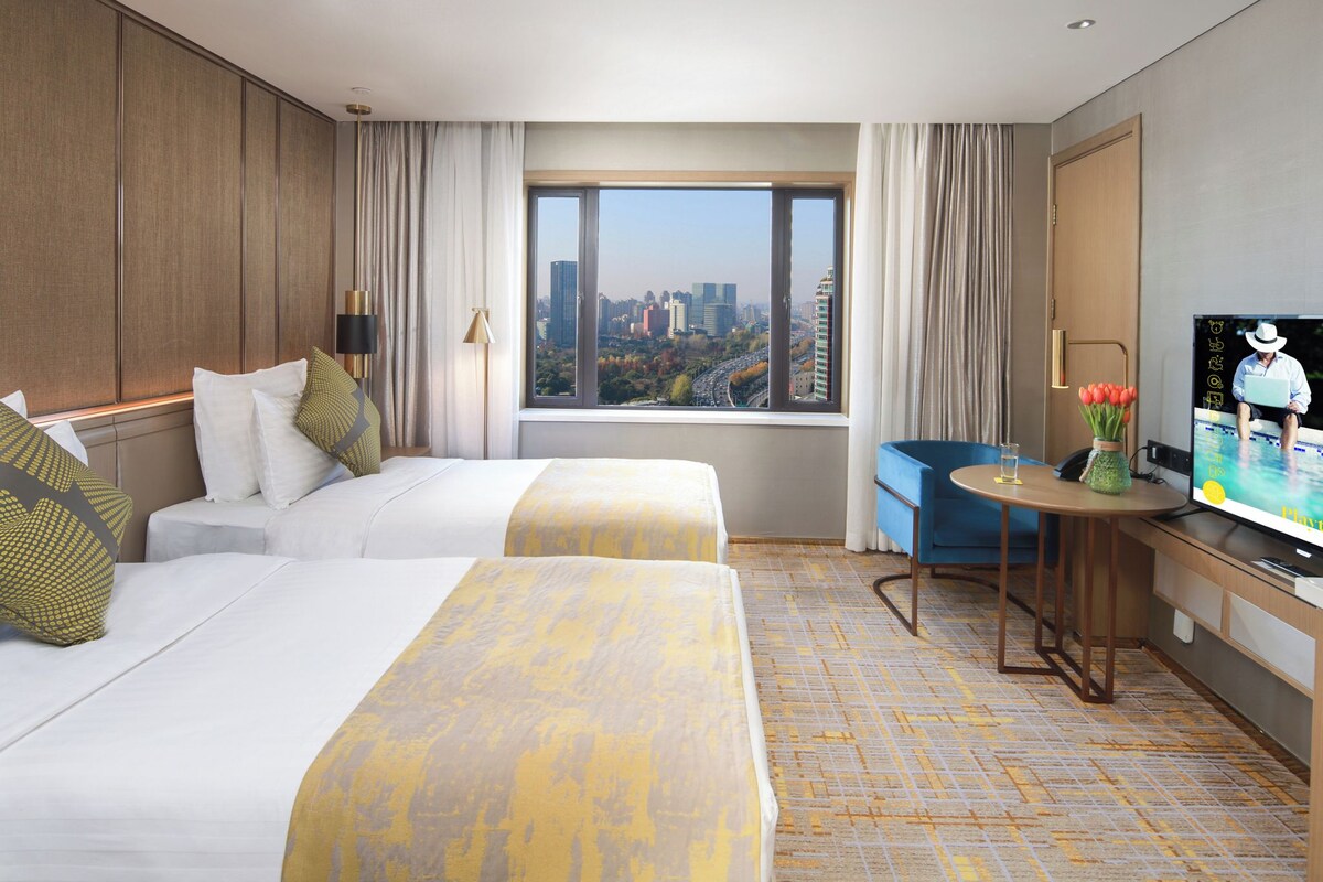 Quarto de hotel com duas camas, uma escrivaninha com televisão, uma janela com vista para a cidade e uma mesinha com um vaso de flores.