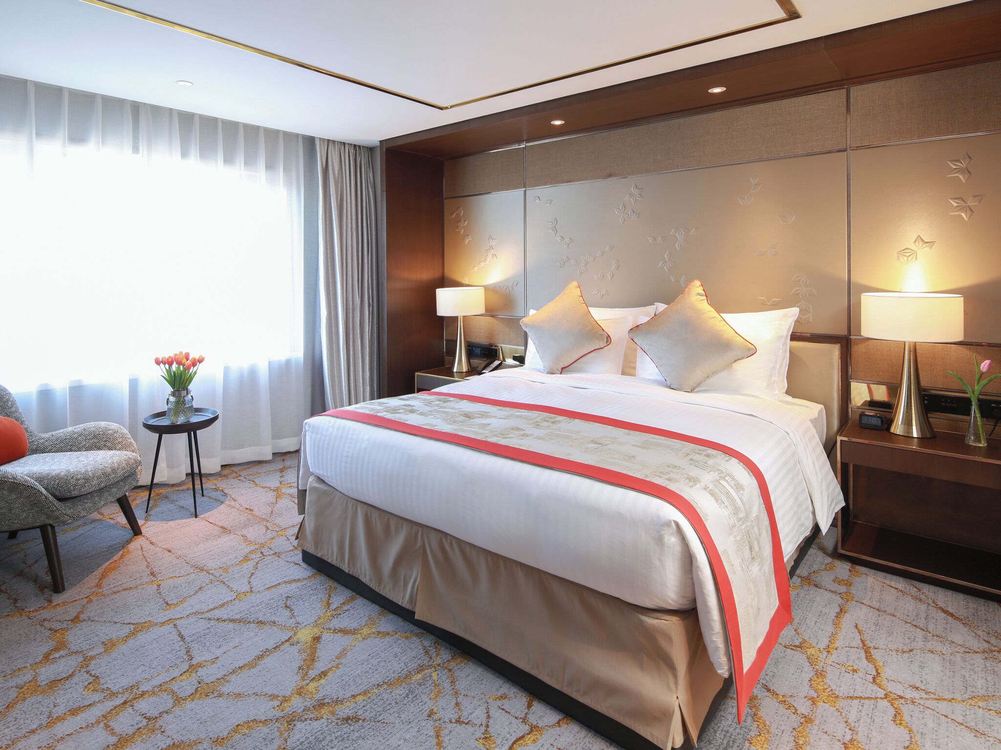 A hotel room with a large bed, two bedside tables with lamps, a small table with a vase of flowers, a chair, and a view of the outdoors through sheer curtains.