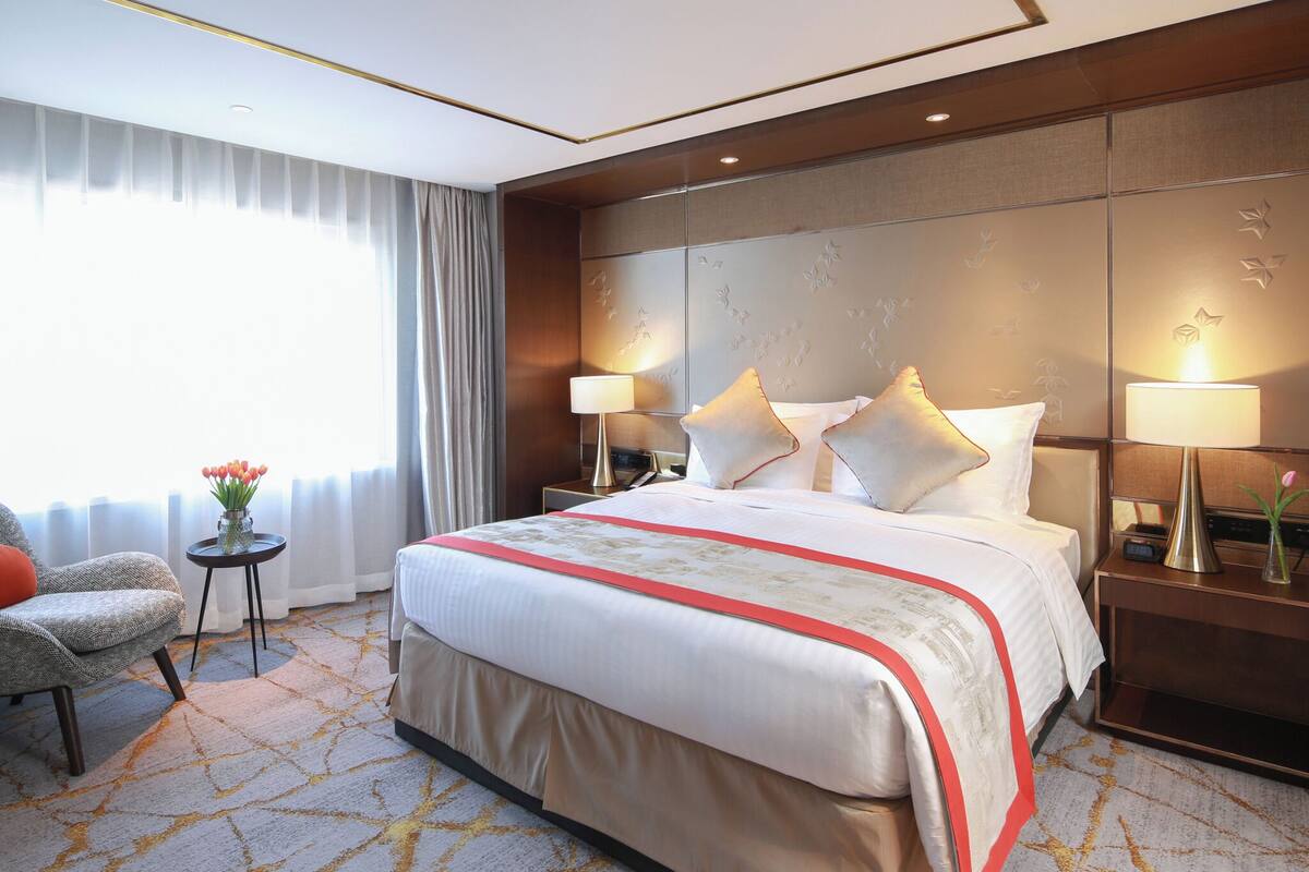 A hotel room with a large bed, two bedside tables with lamps, a small table with a vase of flowers, a chair, and a view of the outdoors through sheer curtains.