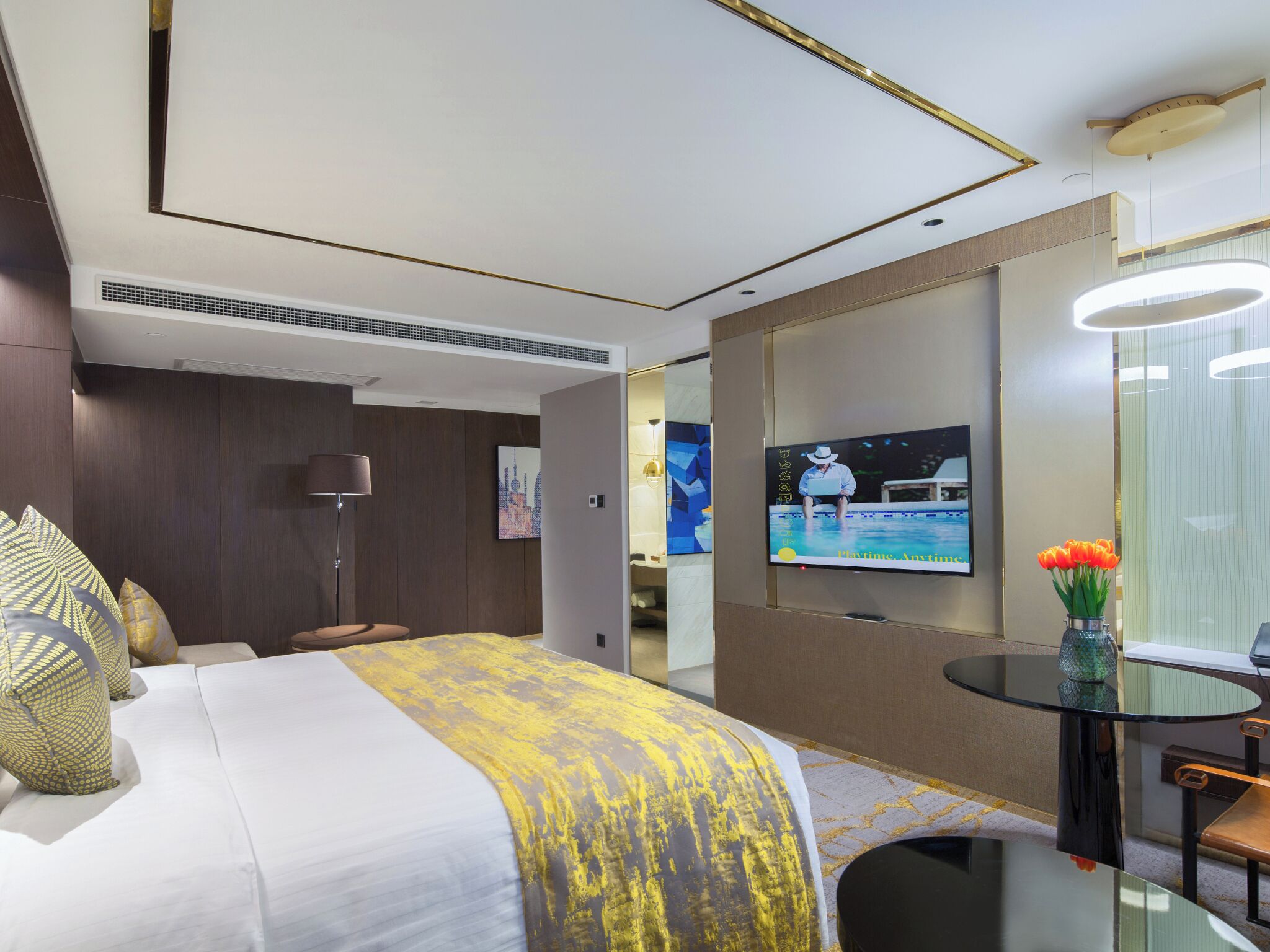 A modern hotel room with a bed, a TV, a desk, and a vase of flowers.