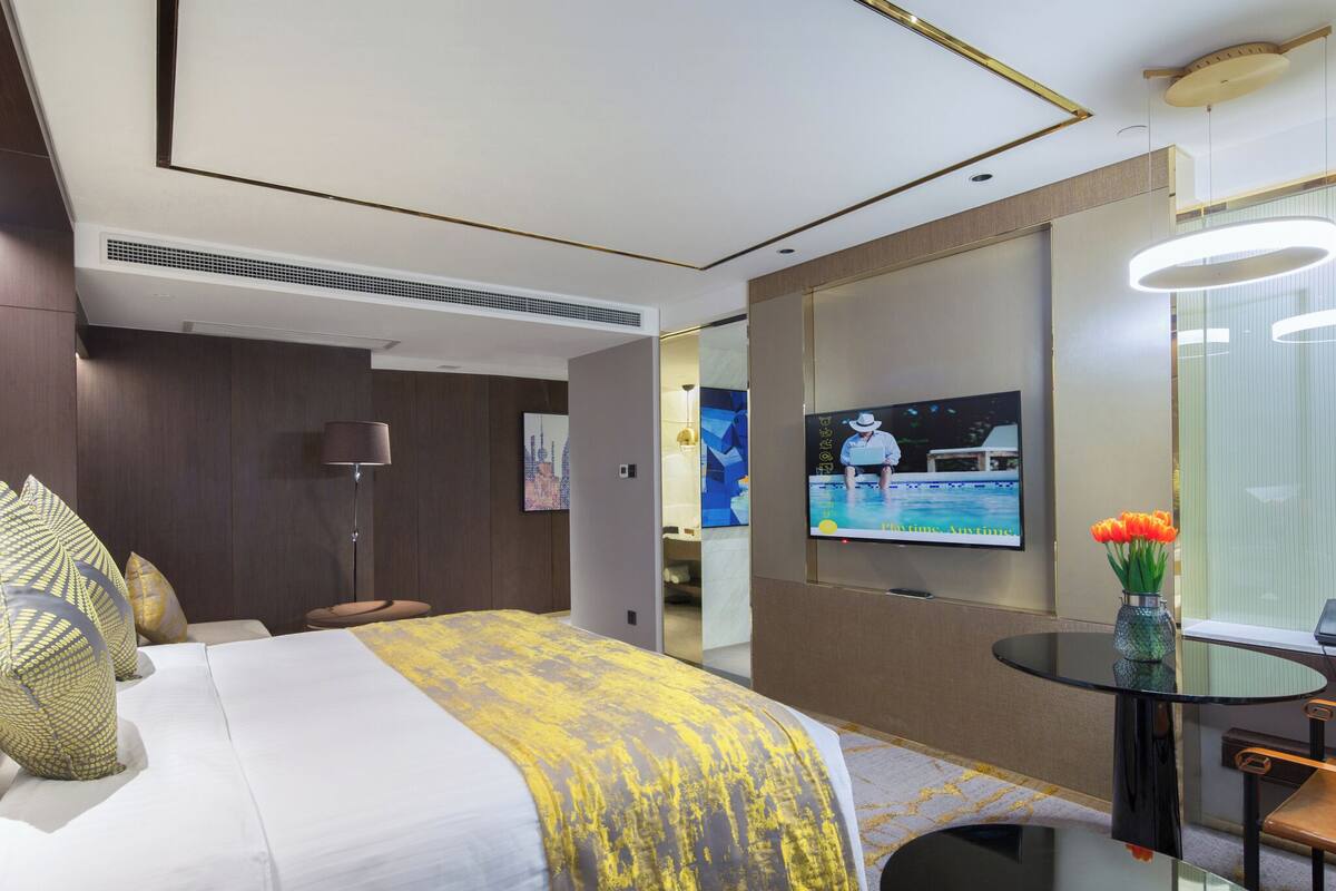 A modern hotel room with a bed, a TV, a desk, and a vase of flowers.