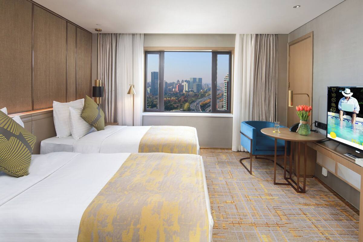 A hotel room with two beds, a desk with a TV, a window with a city view, and a small table with a vase of flowers.