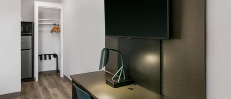 A modern hotel room with a wooden desk, a flat-screen TV mounted on the wall, a microwave, and a mini-fridge.