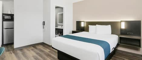 Una habitación de hotel moderna con una cama grande, una mesita de noche, un televisor integrado y un pequeño rincón de descanso.