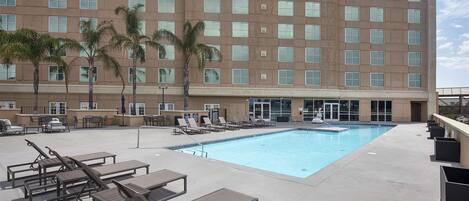 A hotel pool area with sun loungers, palm trees, and a large building in the background.