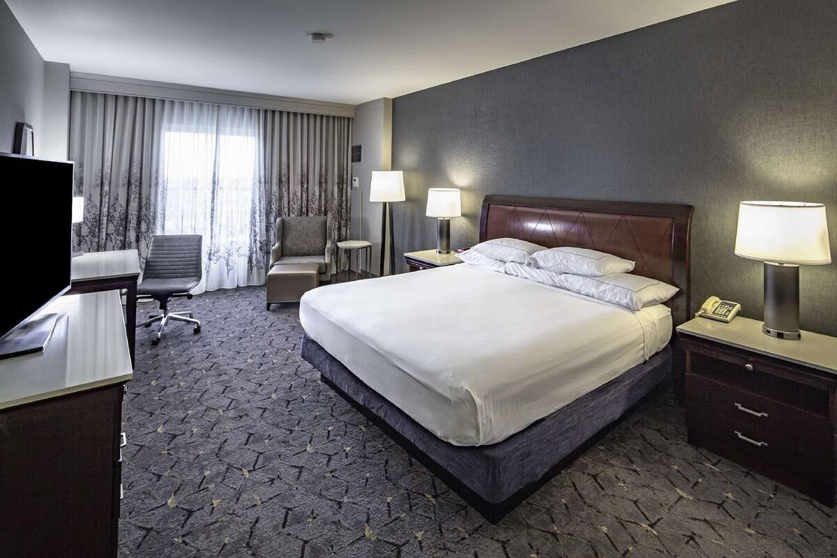 Ett hotellrum med en säng, ett skrivbord, en stol, lampor och en tv.