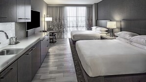 A modern hotel room with a large bed, a desk with a chair, a flat-screen TV mounted on the wall, and a large window with curtains.