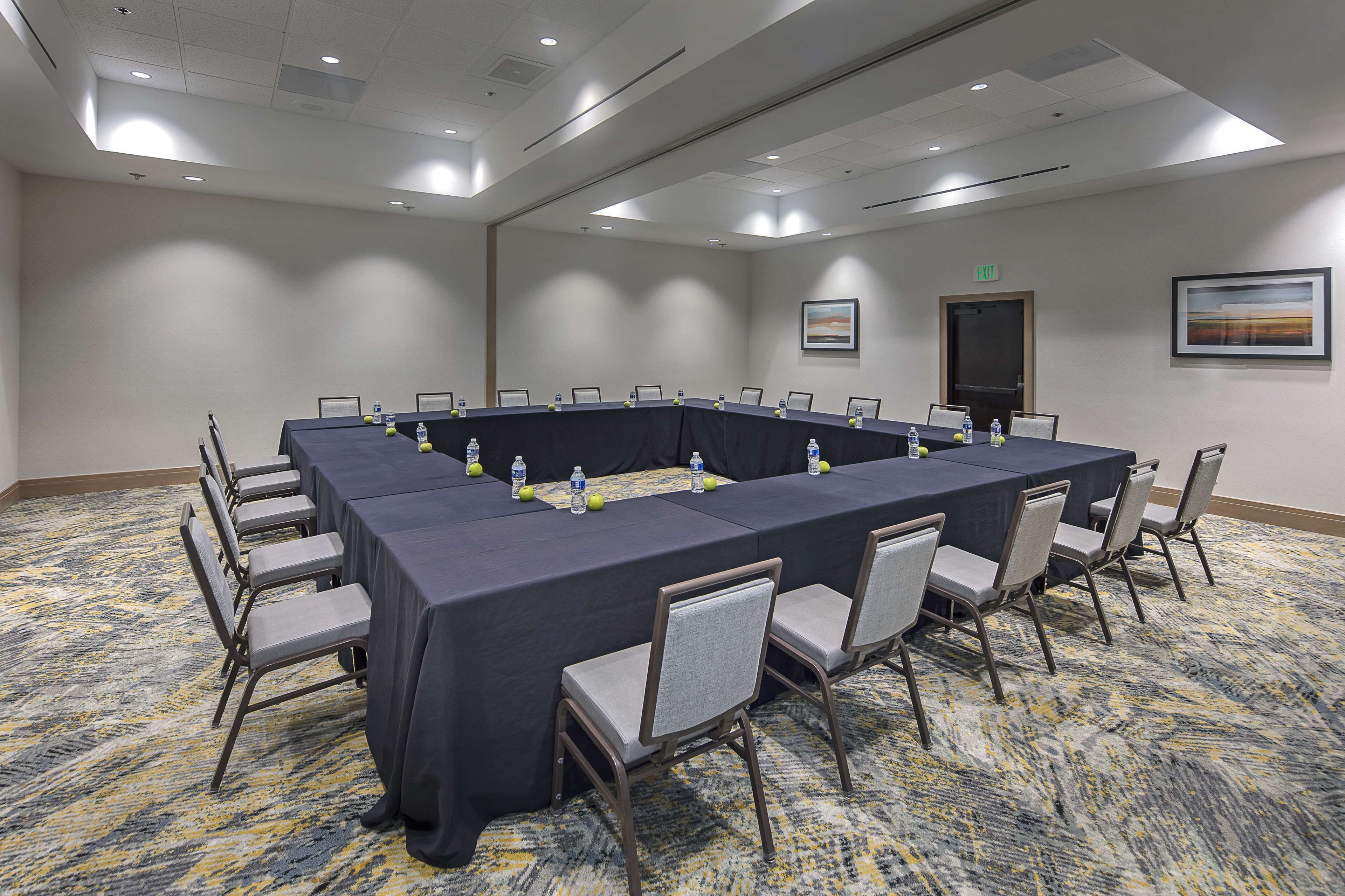Un salón de conferencias con una larga mesa, sillas, botellas de agua y piso alfombrado.