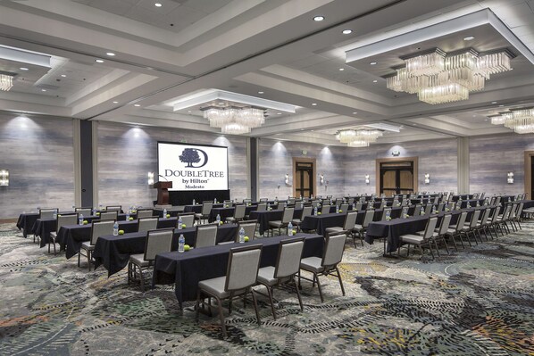 Un salón de conferencias con filas de mesas y sillas, una pantalla grande mostrando 'DOUBLETREE by Hilton Modesto', y candelabros.