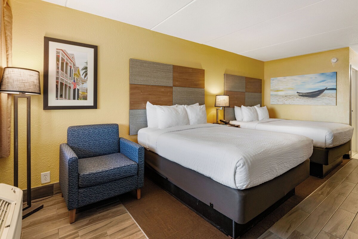 Quarto de hotel com duas camas, uma poltrona azul e um quadro emoldurado de um edifício.