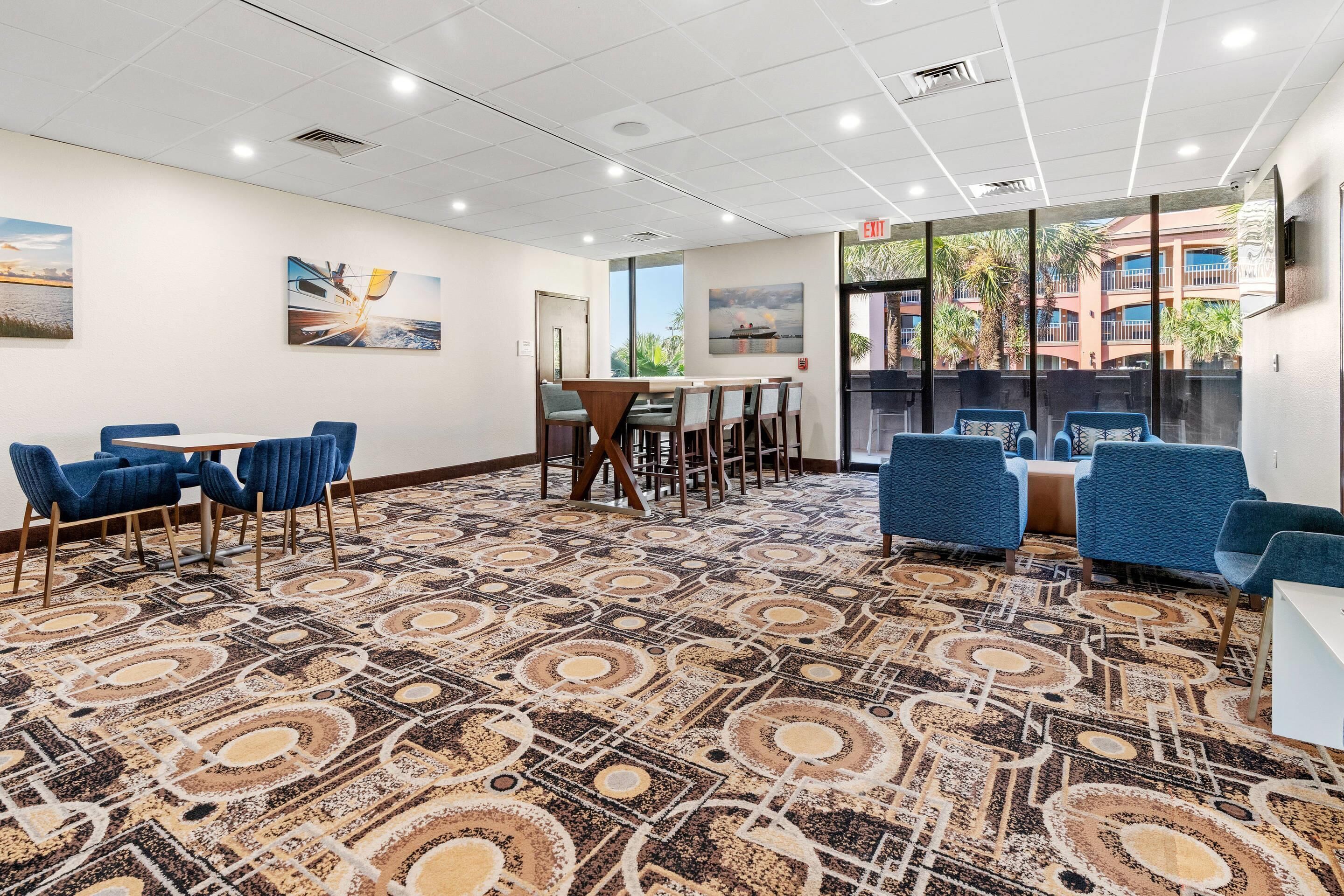 A conference room with a patterned carpet, blue chairs, a wooden table, and a large window with a view of buildings and palm trees.