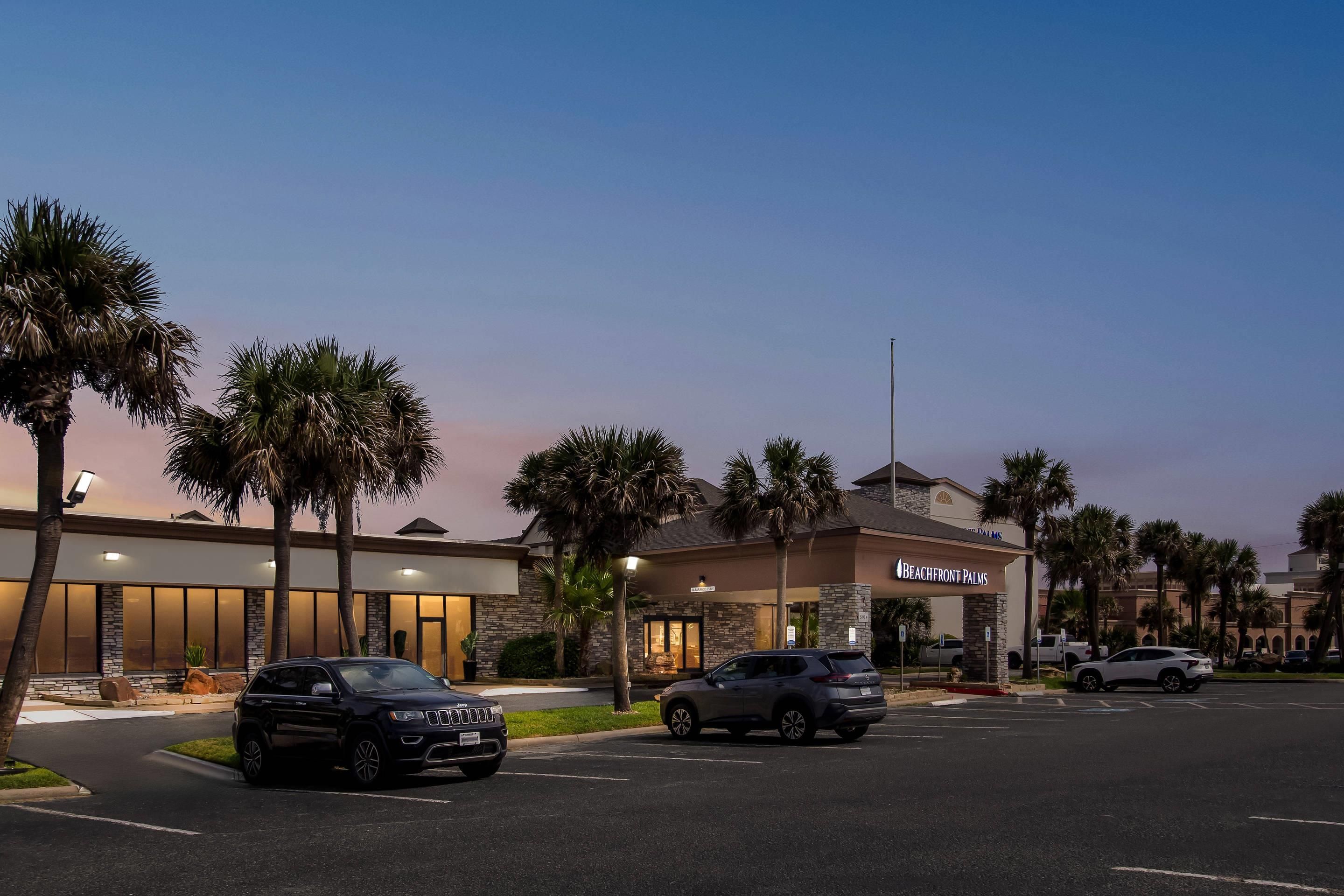 Un hotel con entrada despejada, palmeras y coches aparcados.