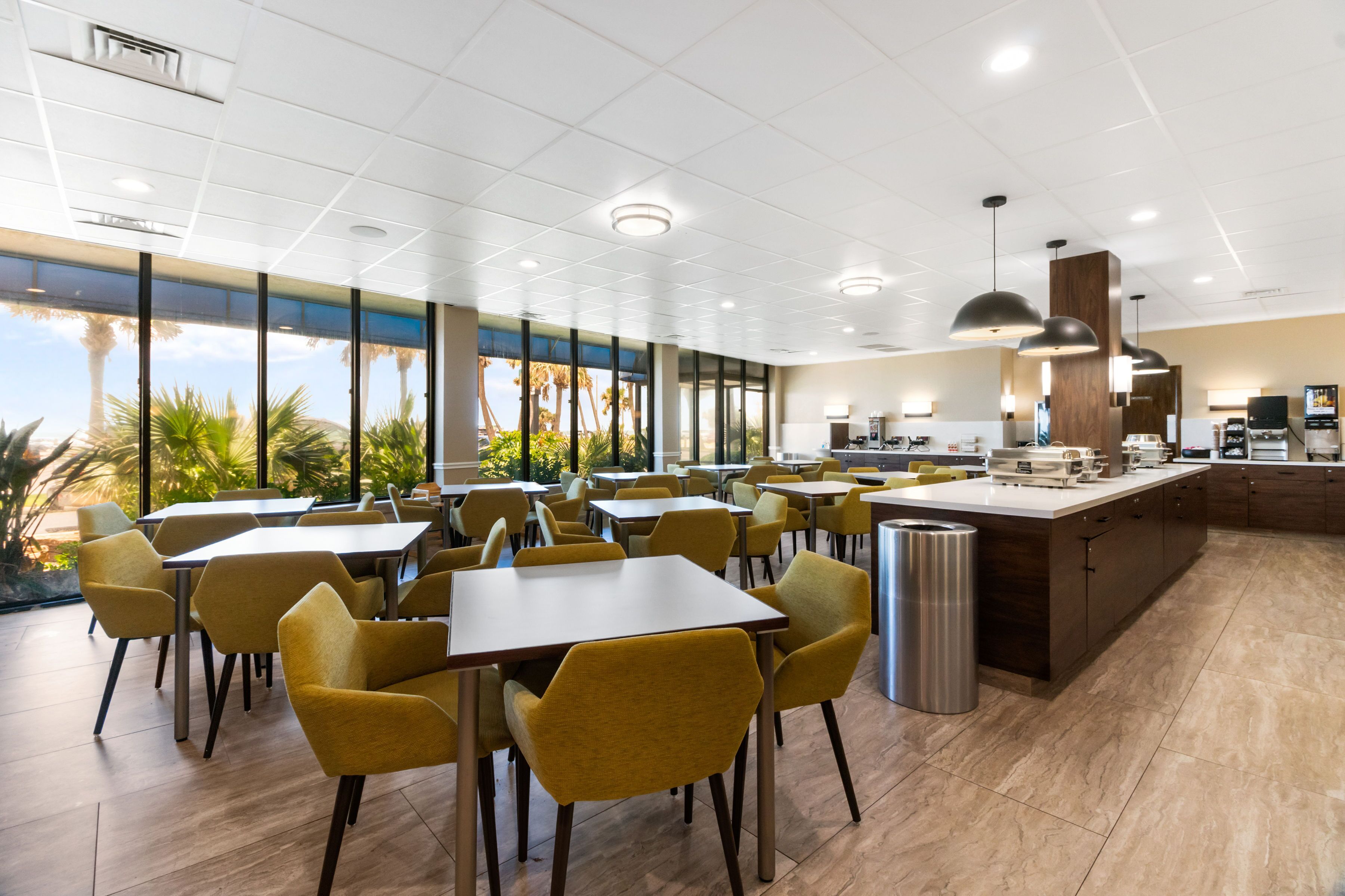 Un restaurant moderne avec des chaises et des tables jaunes, de grandes fenêtres offrant une vue dégagée et un espace cuisine doté d’un comptoir en bois.