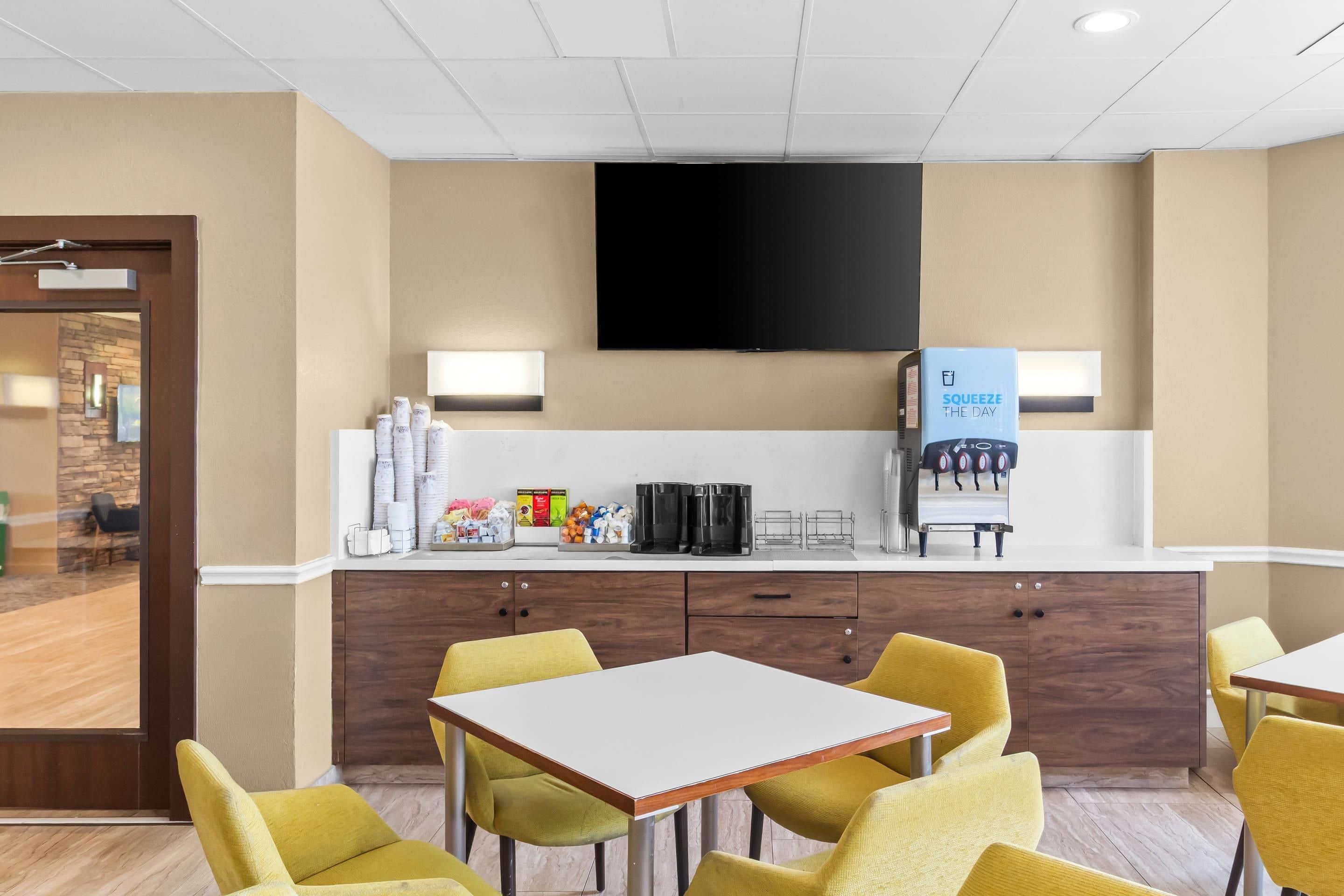 A hotel room with a kitchenette, a flat-screen TV mounted on the wall, a water dispenser, and yellow chairs around a small table.
