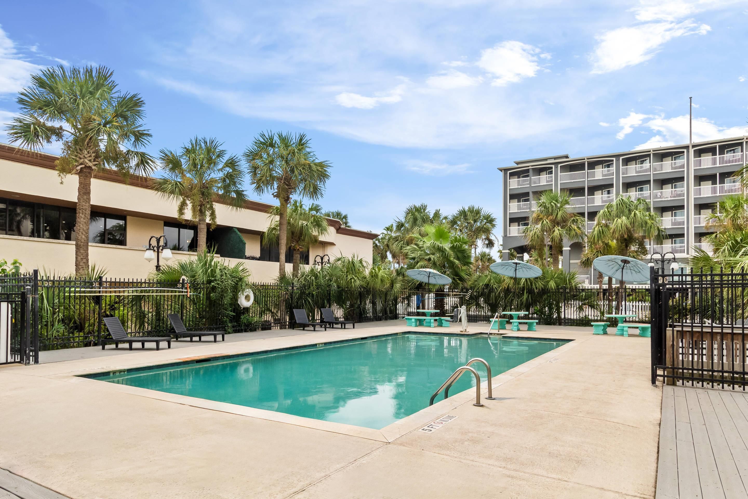 A swimming pool with lounge chairs and umbrellas, surrounded by palm trees and a multi-story building.