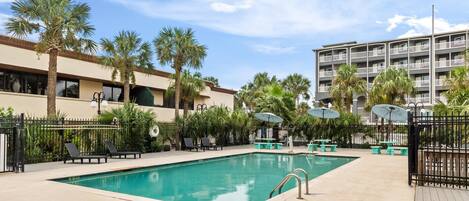 A swimming pool with lounge chairs and umbrellas, surrounded by palm trees and a multi-story building.