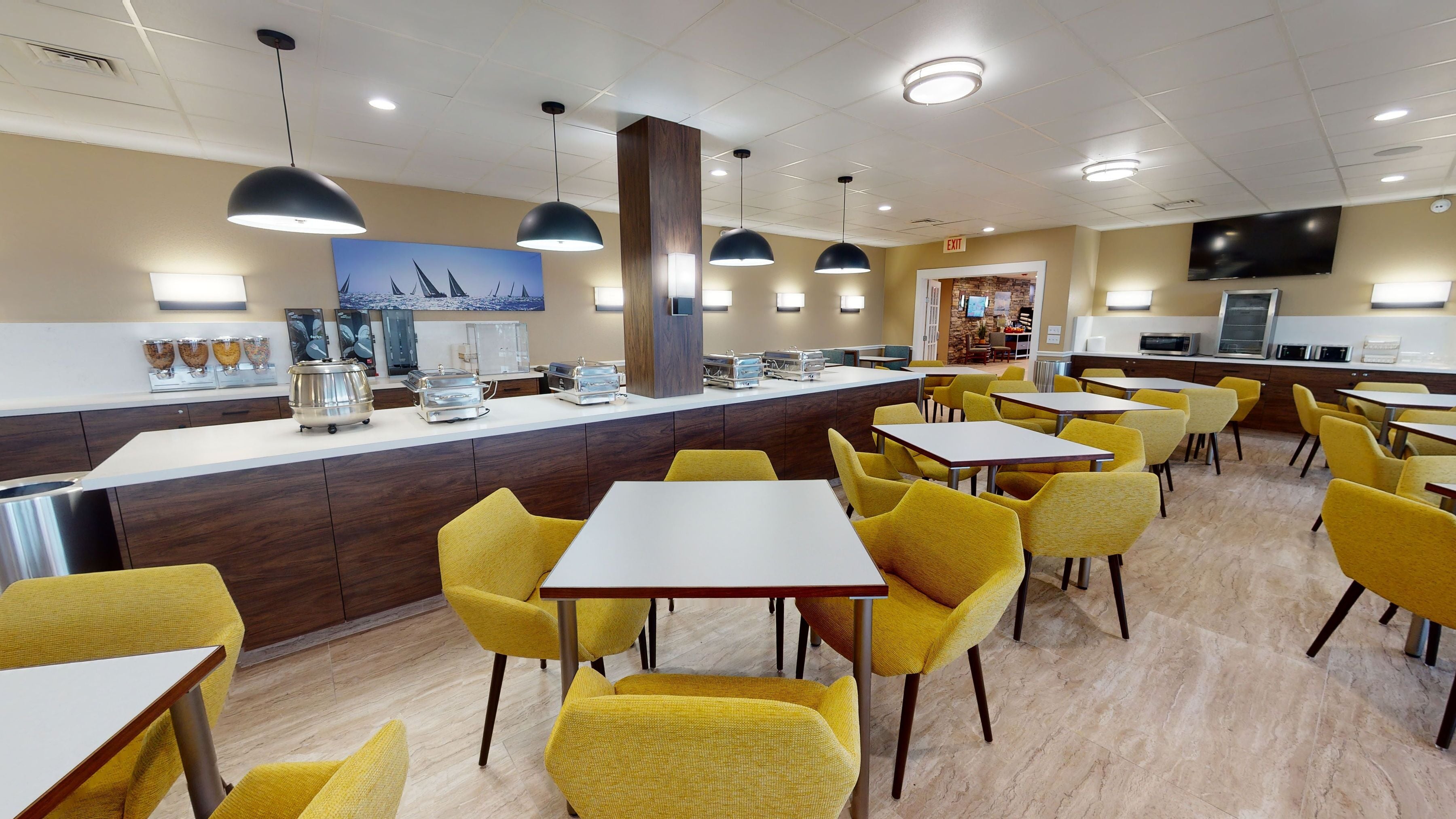 A modern dining area with yellow chairs and tables, a counter with food dispensers, and a TV mounted on the wall.