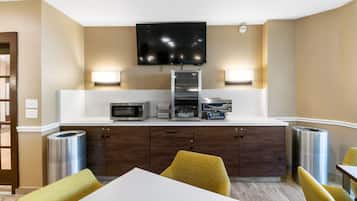 A hotel room with a kitchenette, a flat-screen TV mounted on the wall, and a dining area with yellow chairs.