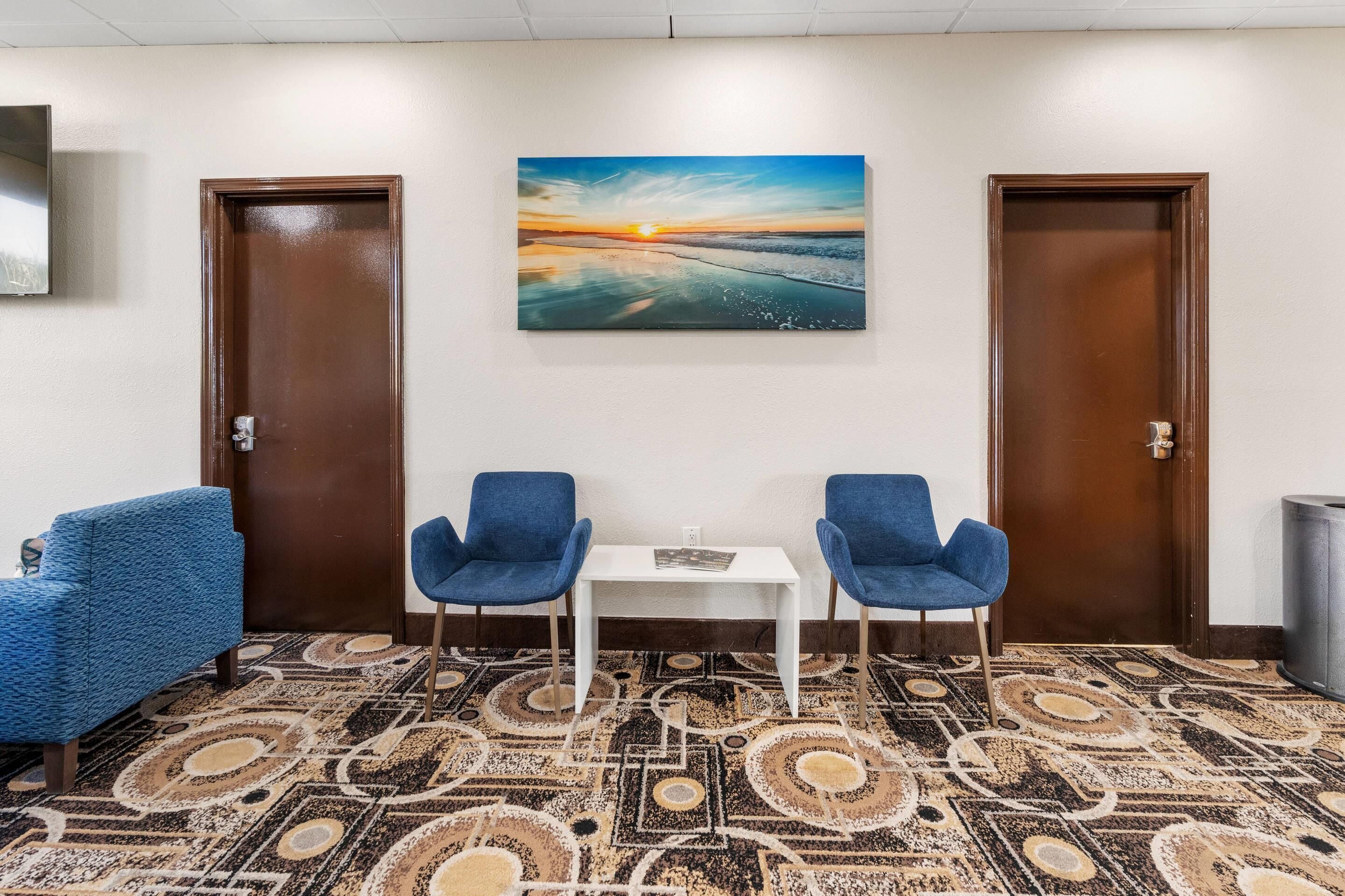 Habitació d'hotel amb una catifes estampada, dues cadires blaves, una taula petita i un quadre amb un paisatge de ponent sobre l'oceà a la paret.