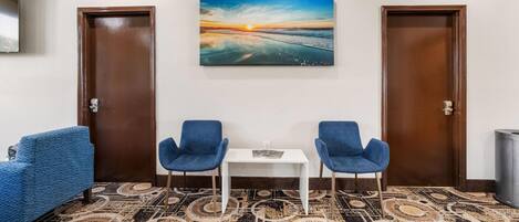 Habitación de hotel con alfombra estampada, dos sillas azules, una mesita y un cuadro con atardecer sobre el océano en la pared.