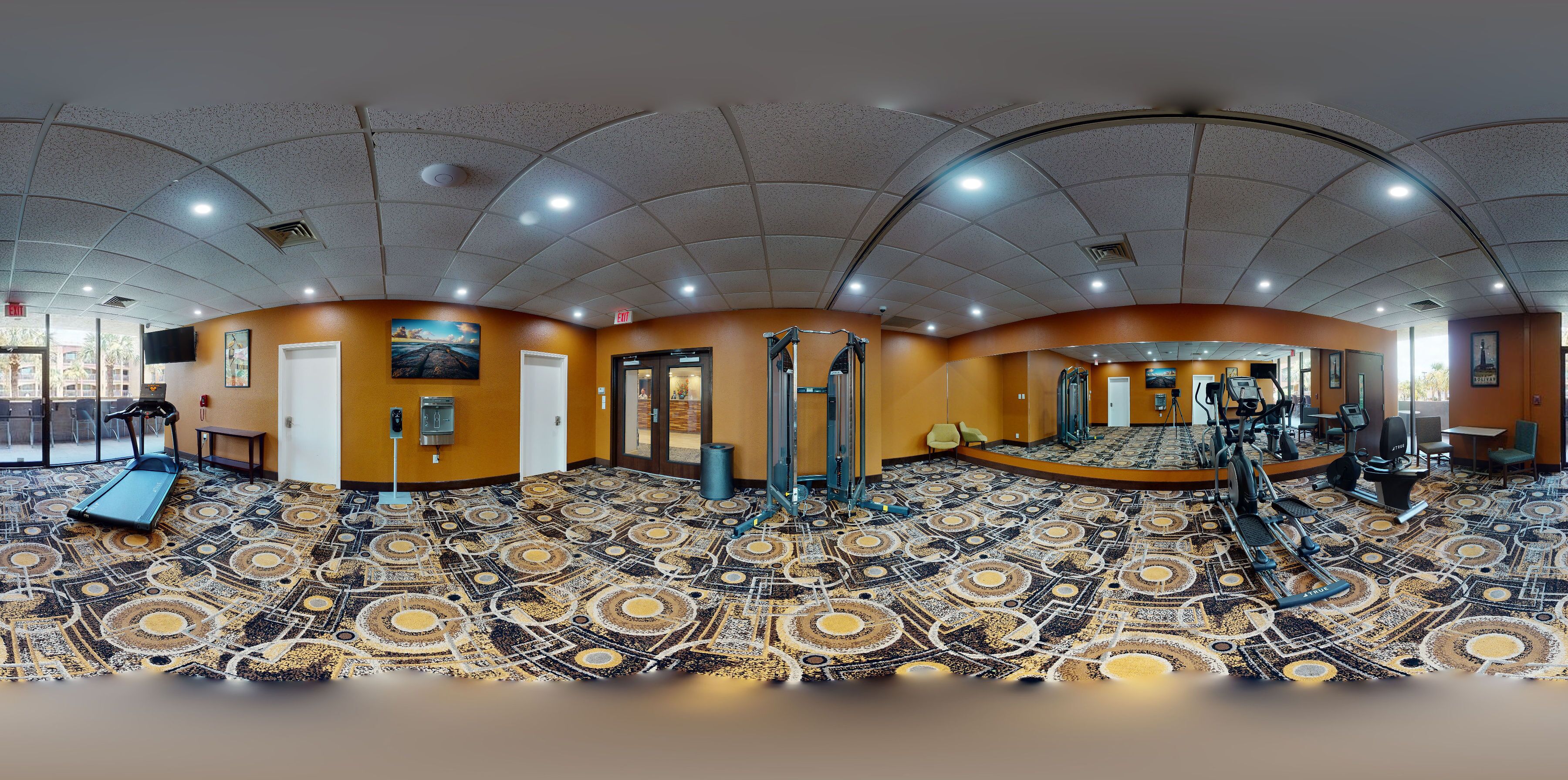 A panoramic view of a gym with exercise equipment, a patterned floor, and orange walls.