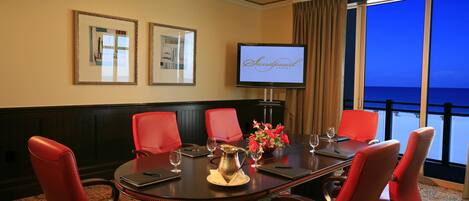 Un salón de conferencias con una mesa grande, sillas rojas, un televisor mostrando 'Sandpearl Resort' y vista al océano a través de amplias ventanas.