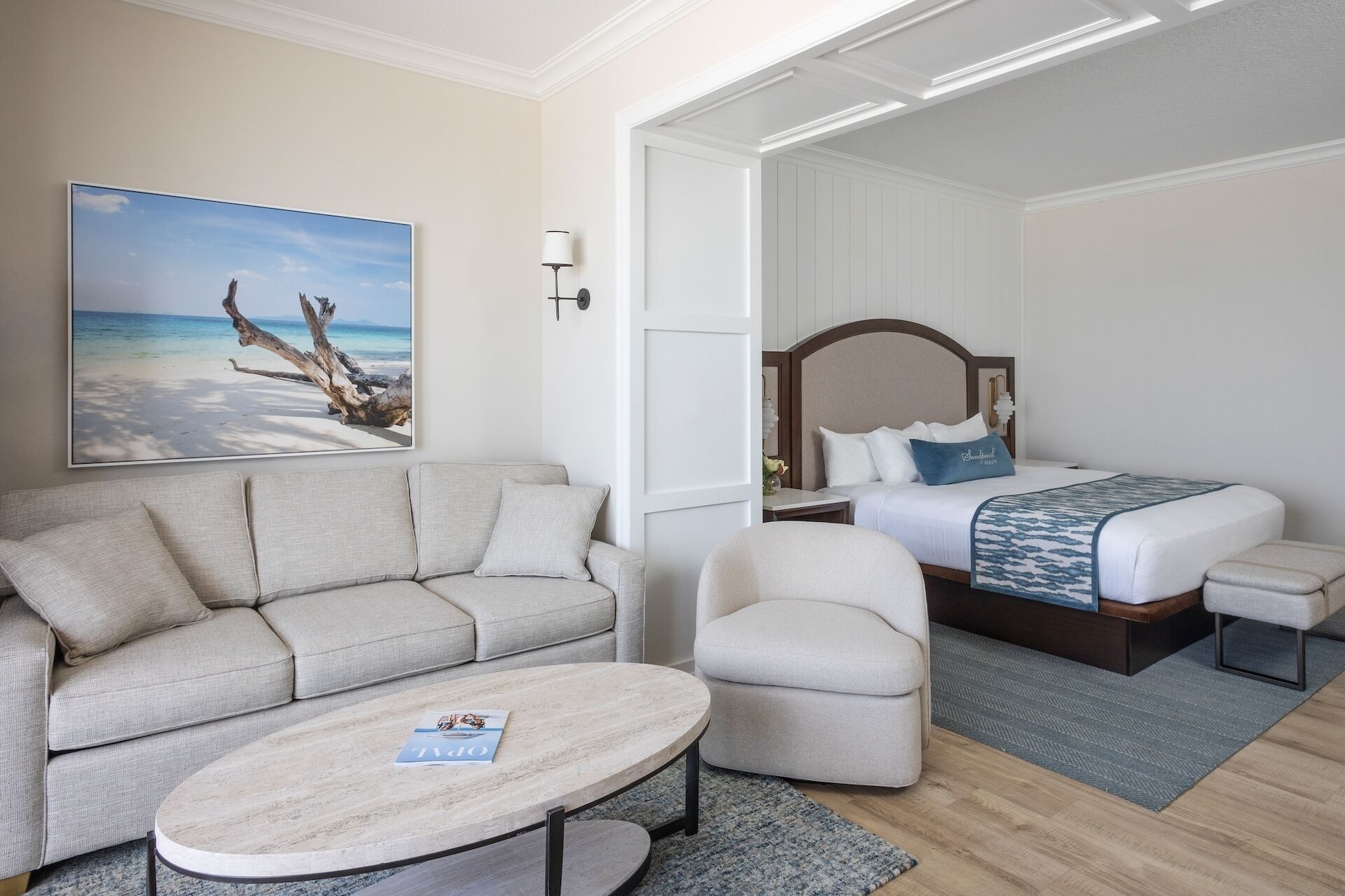 A hotel room with a sofa, a chair, a bed with a headboard, a nightstand, a lamp, and a painting of a beach scene.