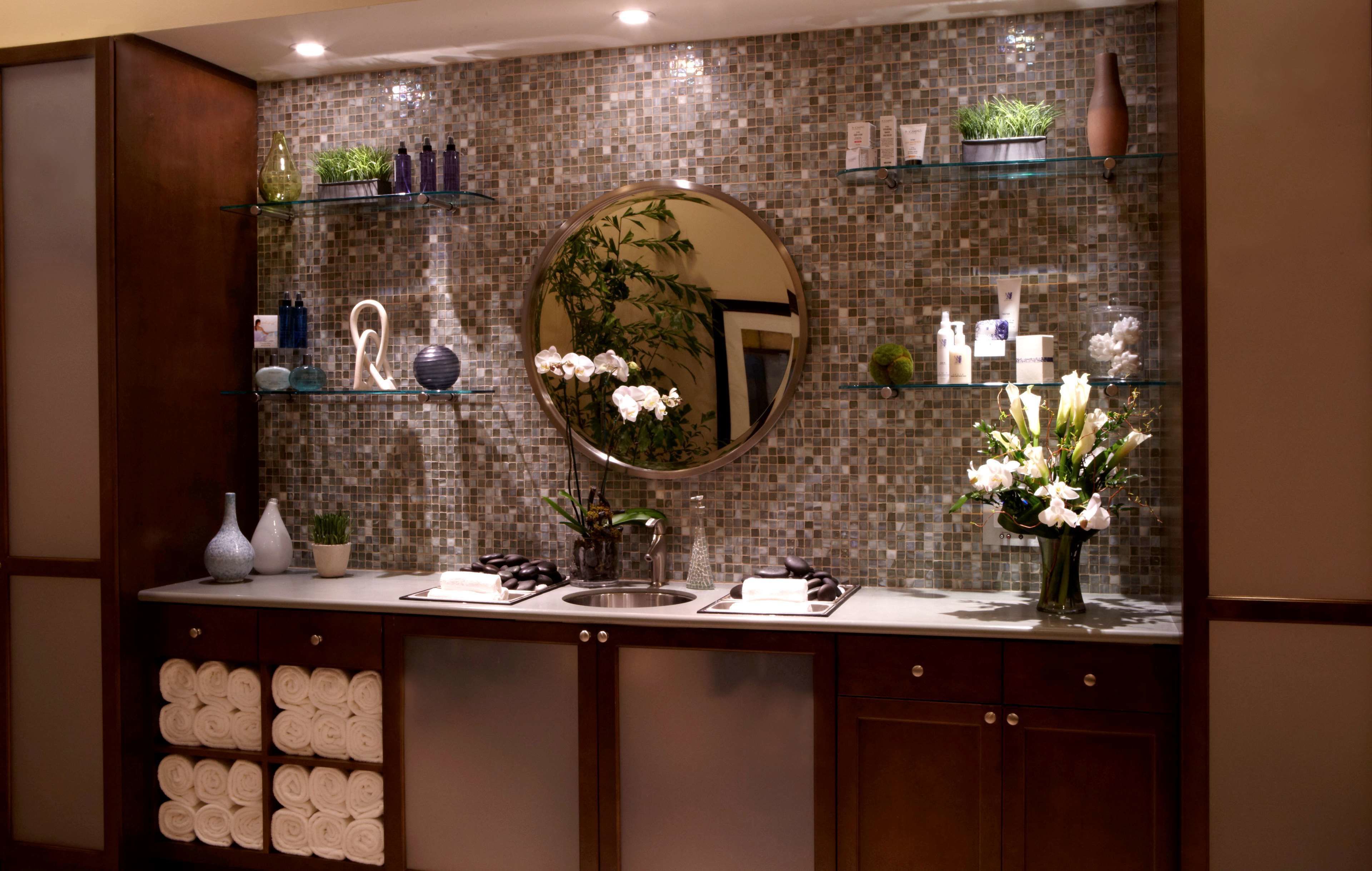 A bathroom with a tiled backsplash, a round mirror, and shelves with toiletries and plants.