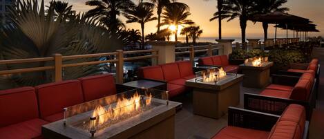 Un lounge en la azotea con asientos rojos, fogones y palmeras al atardecer.