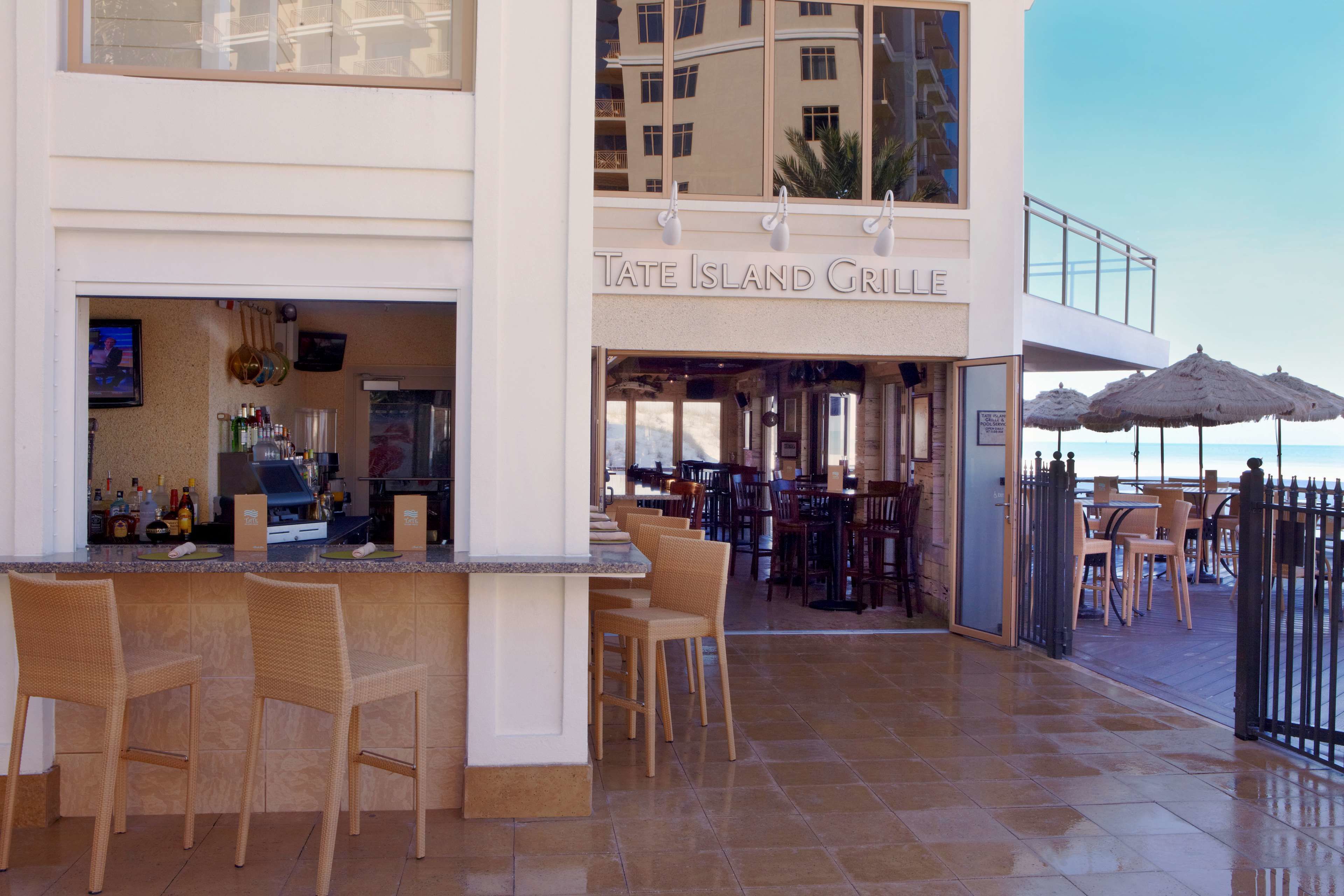 A bar area with wicker chairs and a counter, a dining area with tables and chairs, and a view of the sea.