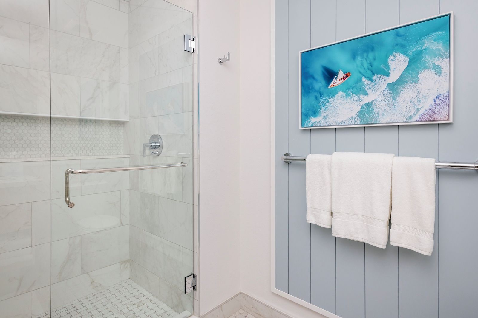 Un baño con cabina de ducha de vidrio, toallero blanco con toallas y un cuadro con una imagen de un velero en la pared.