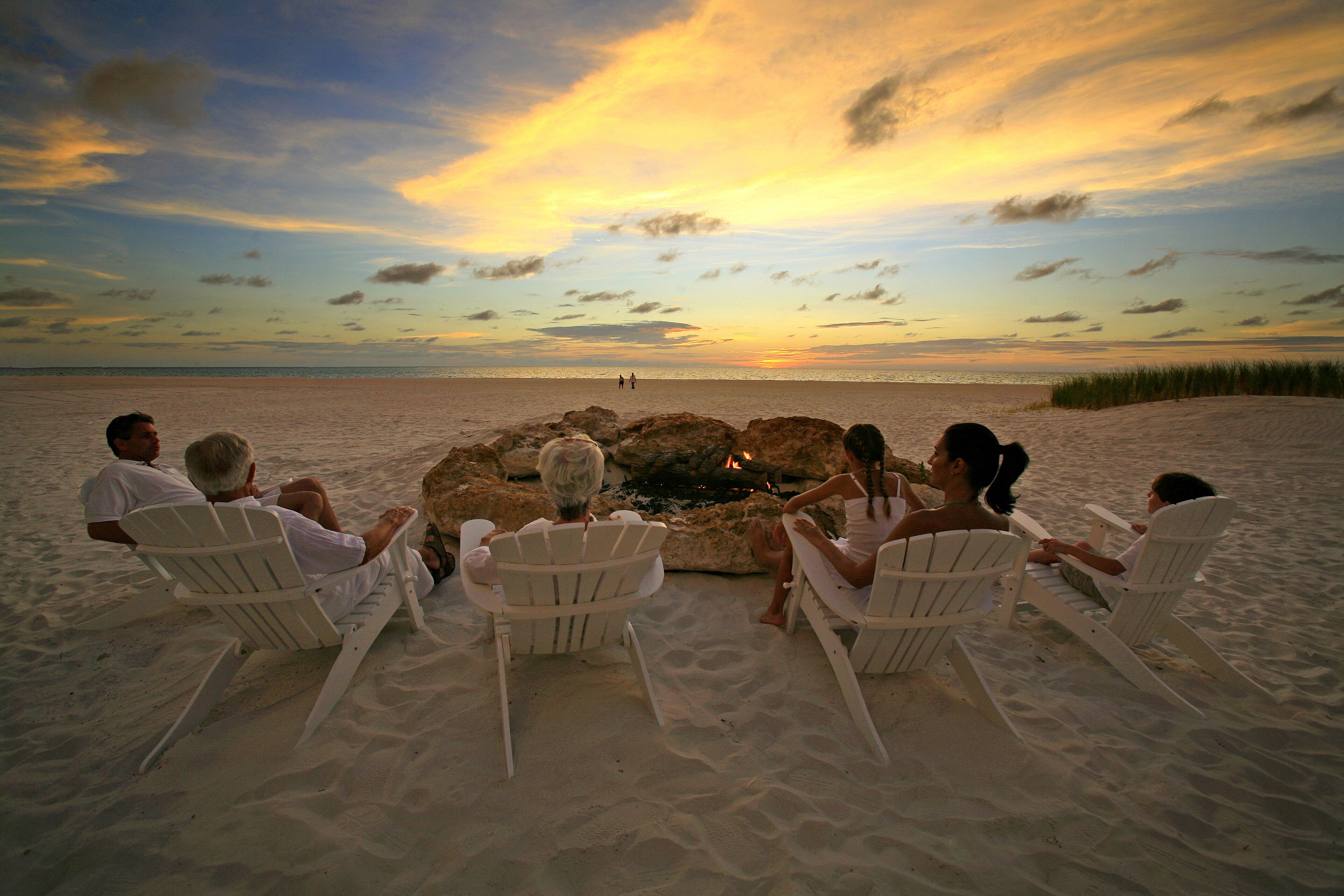 Un groupe de personnes est assis sur des chaises blanches, réparties autour d’un foyer installé sur une plage de sable, avec un coucher de soleil en arrière-plan.