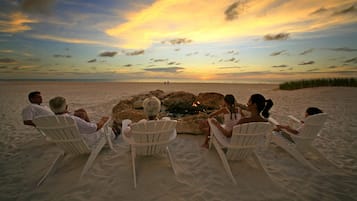 Een groep mensen zit op witte stoelen rond een vuurkuil op een zandstrand, met een zonsondergang op de achtergrond.