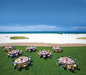 A beachside event with round tables set up on a grassy area, overlooking a sandy beach and the ocean.
