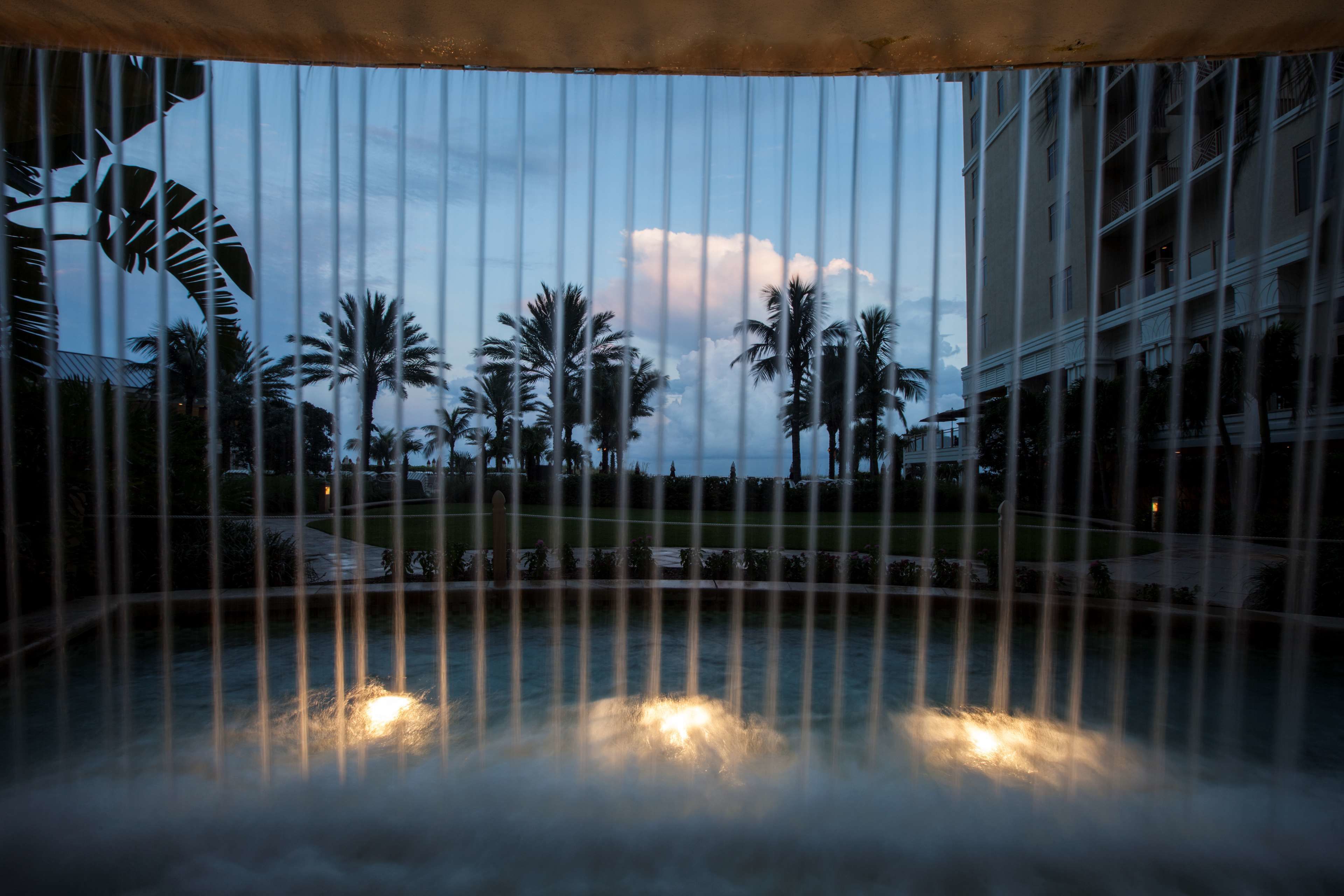 A view through a metal fence of a fountain with water jets, palm trees, and a building in the background.