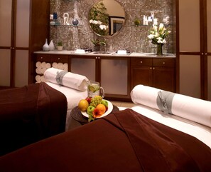 A hotel room with a bed, a bedside table with a fruit plate, a mirror, and a shelf with toiletries.
