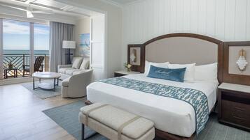 Quarto de hotel com uma cama grande, área de estar com sofá e poltronas, uma mesinha e varanda com vista para o oceano.