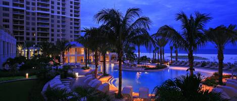 Zona de piscina de un resort con sillones de descanso y palmeras, rodeada por un gran edificio de hotel.