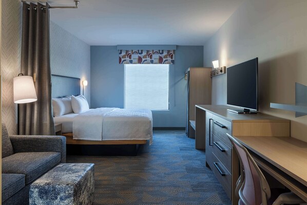 Habitación de hotel con cama, escritorio, televisor, silla y ventana con cortinas.