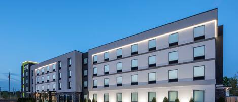 Un hotel moderno con varios pisos, amplias ventanas y un estacionamiento frente a él.
