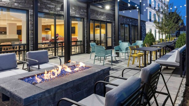 Un patio al aire libre con una chimenea, asientos y un edificio de amplios ventanales y fachada de piedra.