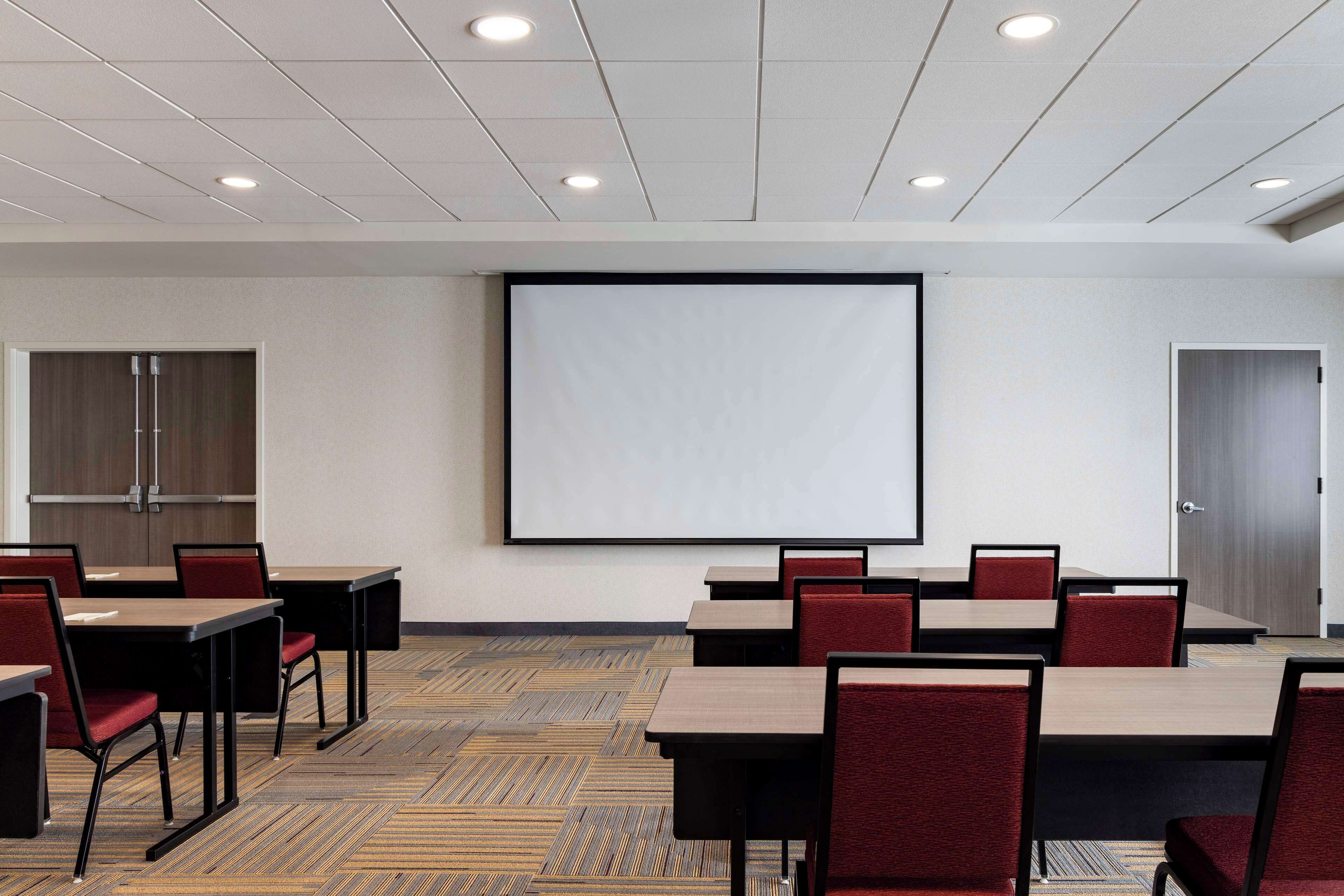 Een vergaderzaal met tafels en stoelen, een scherm voor een projector en een vloerbedekking.