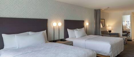 A hotel room with two beds, each with a headboard and bedside lamps. There is a small table and a chair in the room.