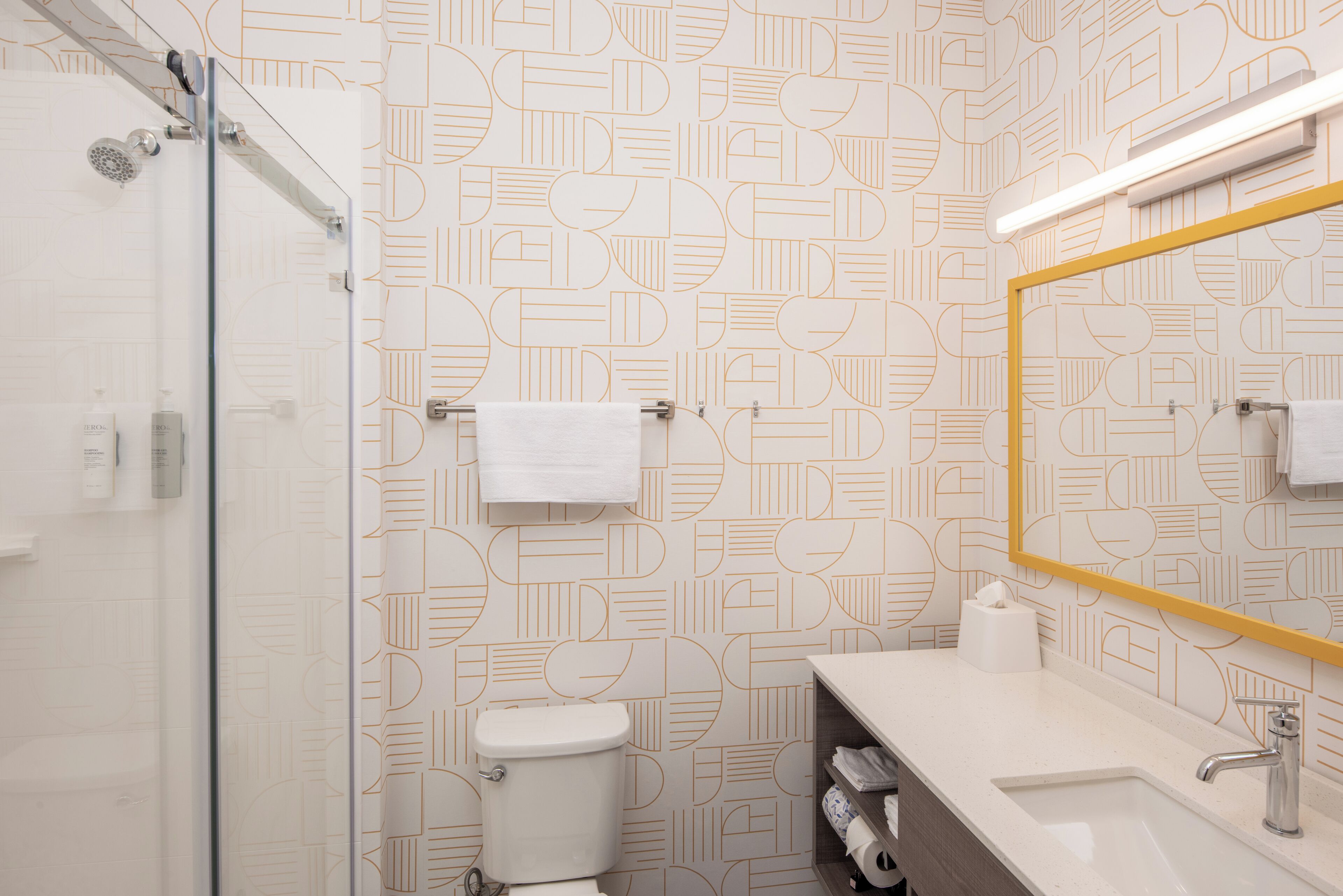 A modern bathroom with a geometric patterned wall, a glass shower enclosure, a white toilet, and a vanity with a mirror and sink.