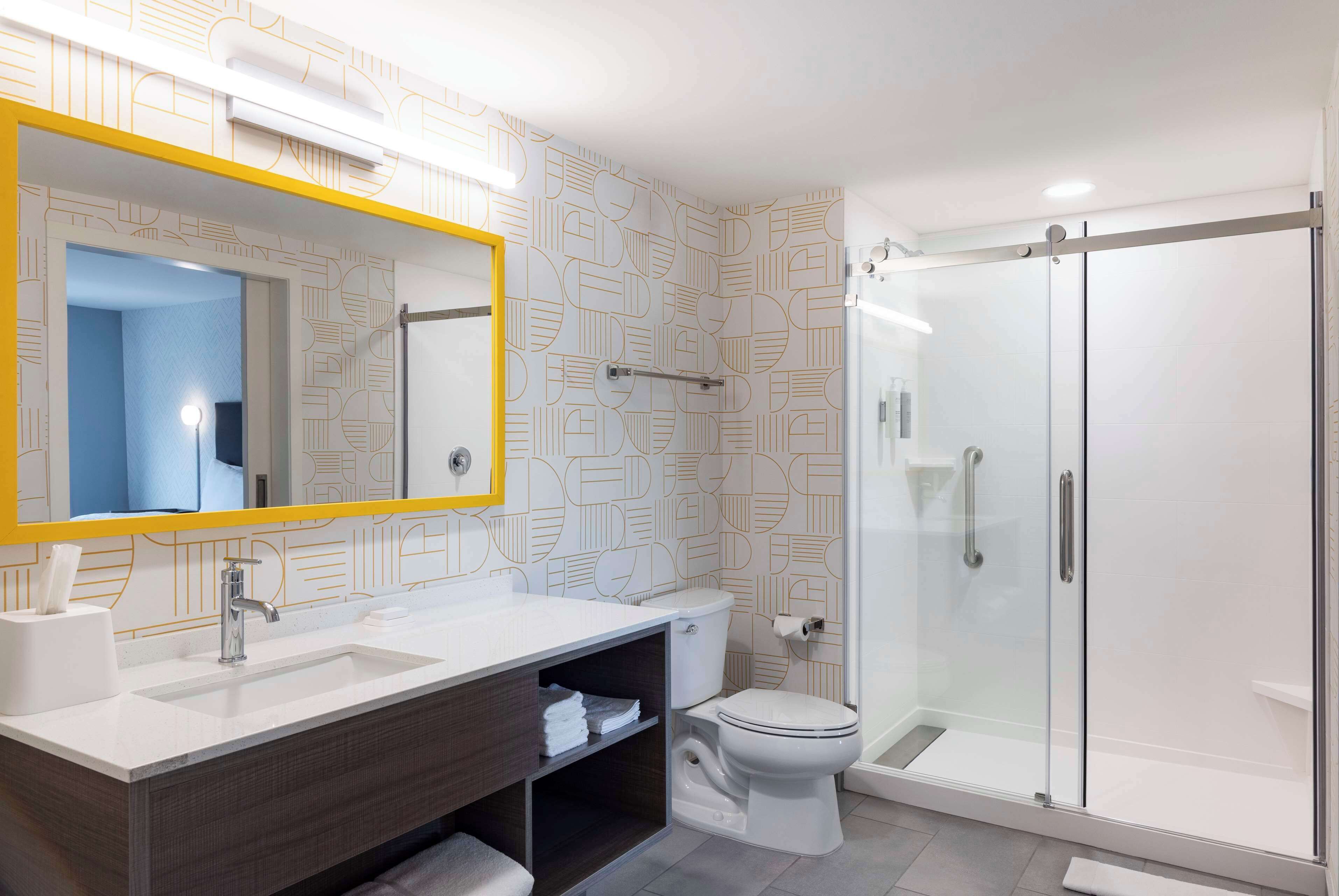 A modern bathroom with a white sink, a mirror with a yellow frame, a toilet, and a glass-enclosed shower.