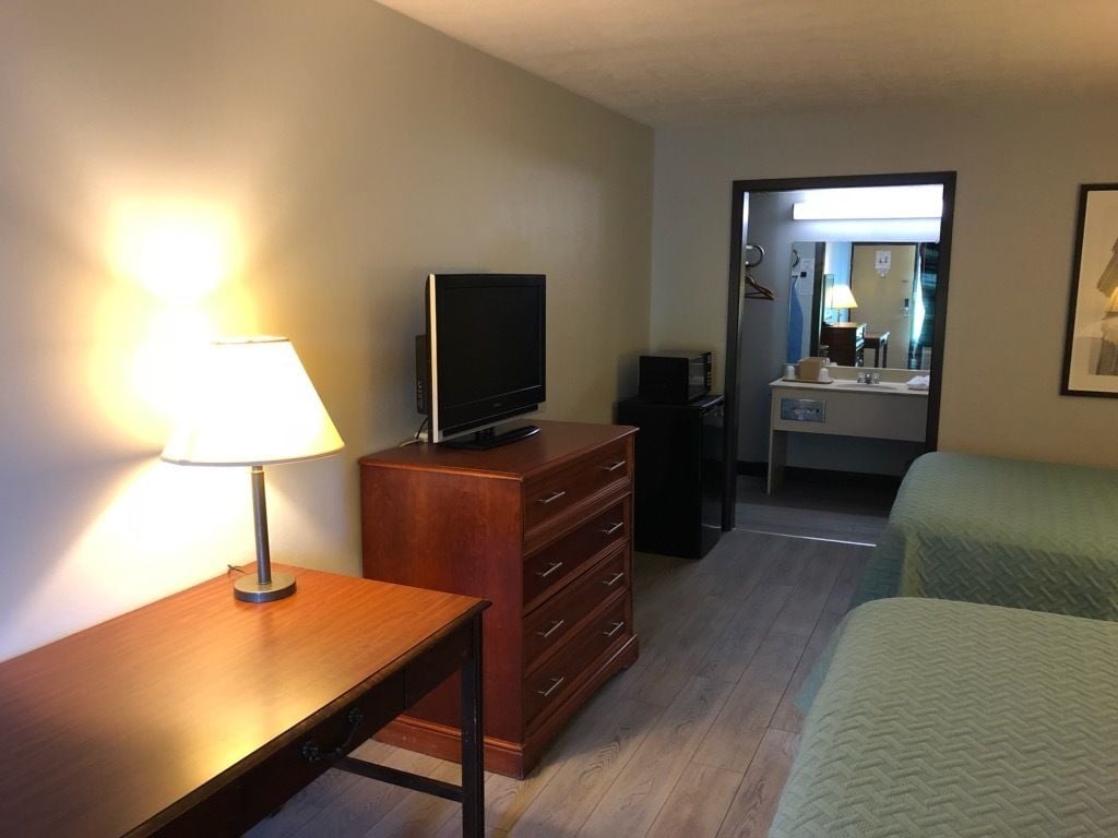 A hotel room with a wooden desk, a television, a dresser, a bed, and a bathroom visible through an open door.