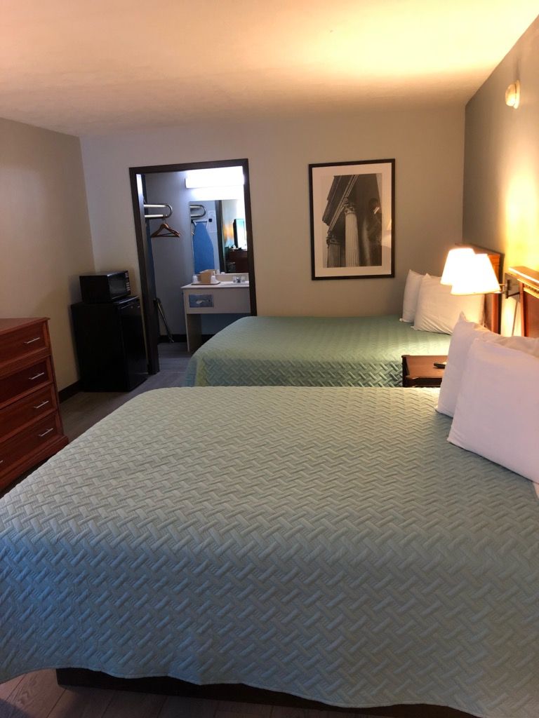 A hotel room with two beds, a dresser, a desk with a computer, a bathroom visible through an open door, and a framed picture on the wall.