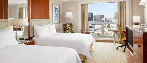 A hotel room with two beds, a desk, a chair, a TV, and a large window with a city view.